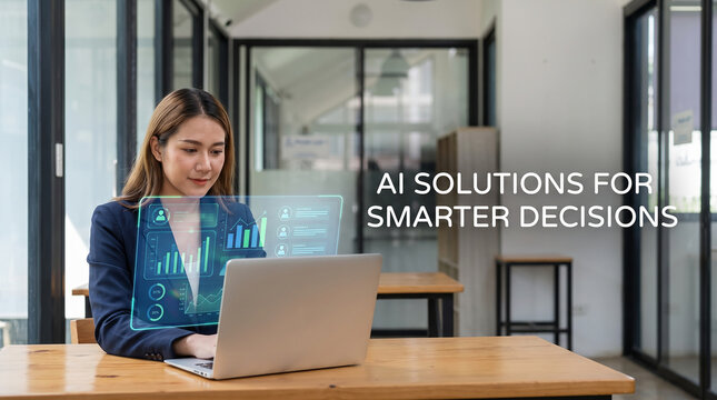 Asian businesswoman interacting with AI assistant hologram displaying financial charts and business data on laptop in a minimalist office setting.