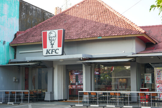 Exterior shot of a famous KFC fast food restaurant building outlet showcasing the recognizable Colonel Sanders logo signboard, front glass entrance doors, tiled red roof, and empty outdoor chairs.
