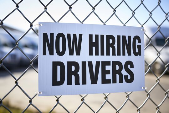 White rectangular sign displaying bold black text now hiring drivers attached to a chain link fence with blurred vehicles in background.