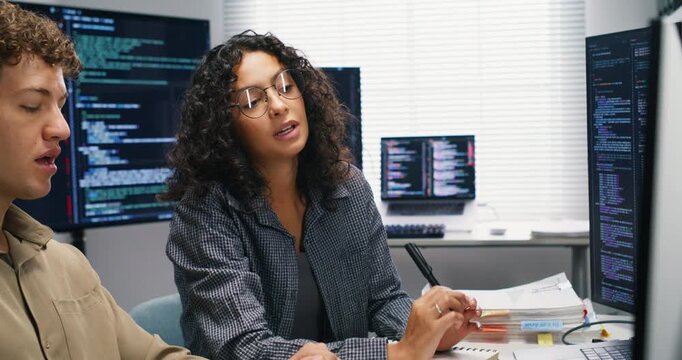 Two Hispanic software engineers pair-program AI models at a shared workstation, reviewing datasets and model metrics on multiple monitors. They discuss optimization strategies, version control, and de