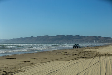 Convertible off-road vehicle driving along a sandy ocean beach on a summer evening, with hazy rolling hills extending across the distant background. The scenic coastal setting combines rugged driving © Val