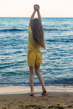 Young girl with long hair standing barefoot on sandy shore, arms raised, facing the ocean on a bright summer day.