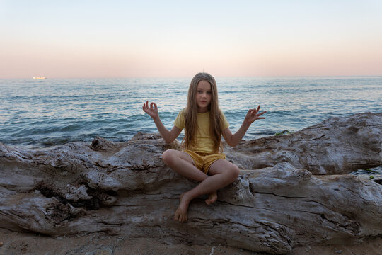 Young girl in yellow sitting cross-legged on driftwood by the sea at sunset, practicing meditation in a peaceful beach setting.