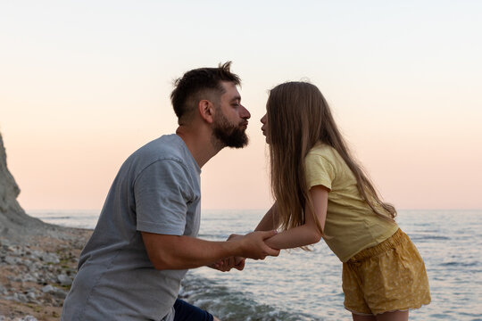 Man and young girl hold hands, sharing a tender moment by the seaside at sunset, with soft pastel sky and calm waves.