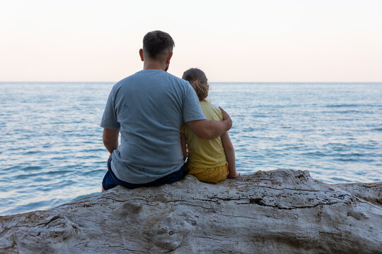 Rear view of father and daughter sitting on driftwood, looking at the calm sea under a clear sky, evoking family bonding and peaceful moments.