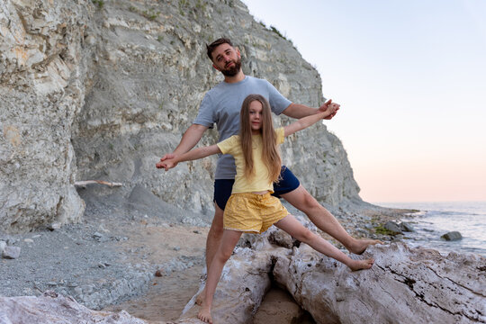 Father and daughter balancing on driftwood at a rocky beach, with cliffs and soft sunset light, conveying family bonding and outdoor adventure.