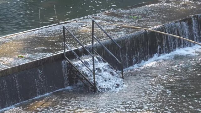 A black metal staircase submerged in water at a small river dam with flowing rapids.