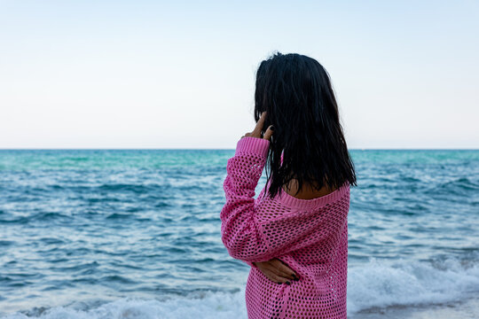 Woman in a pink sweater stands by the sea, gazing at the waves. Serene beach scene with calm water and clear sky.