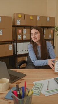 Young woman holds shipping box with both hands and gestures toward laptop in an office building; focused entrepreneurship.