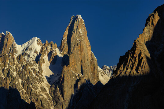 View of the Uli Biaho granite spire rising against a clear deep blue sky, featuring snow-capped ridges and dramatic rock faces in the Karakoram Range, Gilgit Baltistan, Pakistan.