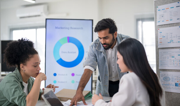 Male team leader leaning over the desk to explain project details to colleagues, presenting a marketing research analysis on a digital display in a modern corporate office.