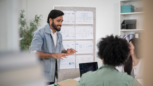 Confident male executive standing and using hand gestures to explain a business strategy to his colleagues during a collaborative meeting in a bright modern office.