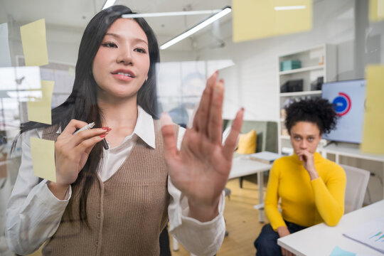 Creative businesswoman brainstorming and writing project strategy on a clear glass board with a marker while her colleague watches carefully.