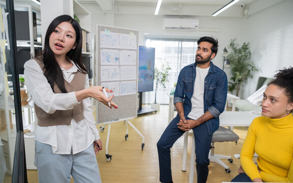 Female team leader standing and explaining a strategic plan to a male colleague who is sitting on the desk listening attentively.