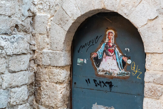 Bari, Italy - June 31, 2026: Street art sticker on weathered metal door in arched stone doorway depicting Virgin Mary figure wearing Superman logo in old town alley