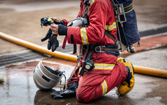 Firefighter Putting On Black Protective Gloves while Kneeling beside a Silver Helmet and Fire Hose on Wet Pavement
