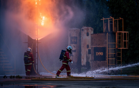 Firefighter Team Moving through Water Spray beside a Burning Industrial Structure while Wearing Protective Uniforms and Air Tanks