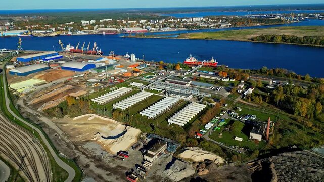 Aerial view of industrial port facilities with storage tanks, railway tracks, and ships docked along the river in Latvia.