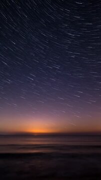 Long exposure star trails over a calm ocean horizon at dusk with purple and orange sky.