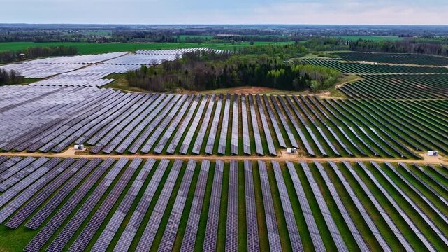 Aerial view of a large solar farm with rows of photovoltaic panels set in a rural landscape with green fields and trees in Latvia.
