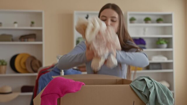 Woman sorting donated clothes, reaching into and then crossing arms beside a cardboard donation box in a building; contemplative giving.