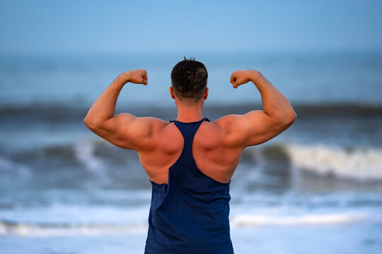Muscular man shows strong back. Beach muscular pose highlights physique. Muscular shoulders show bodybuilding power. Healthy muscular lifestyle supports wellbeing.