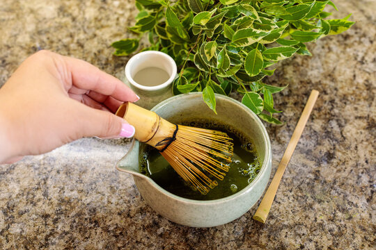 A hand holds a bamboo whisk above a ceramic bowl filled with green matcha liquid, preparing to whisk it smooth