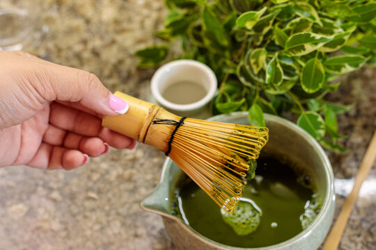 A hand holds a bamboo whisk above a ceramic bowl filled with green matcha liquid, preparing to whisk it smooth