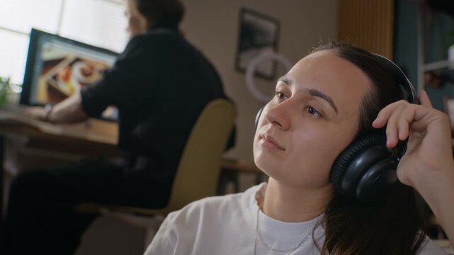 Young Caucasian woman wearing headphones relaxing with eyes closed, taking break from remote work while boyfriend works in background. Perfect for mental health or audio equipment campaigns