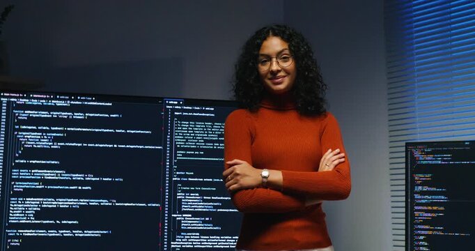 Confidence Hispanic woman at standing with armcrossed in front of computer monitor reviews microservice diagrams and deployment scripts, focused and strategic.