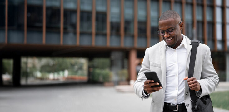 Using smartphone, texting, black businessman outdoor near office building, works as sales manager. African american business man smiling, prepared for client meeting. Copy space. Web banner.