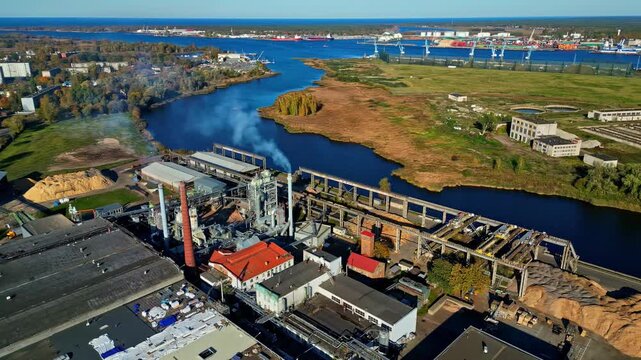 Aerial view of industrial wood processing facility with a tall chimney and wood chip piles along a river in Latvia.