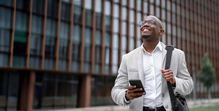 African american businessman working as marketing manager, holding smartphone, looking aside, outdoor near office building. Black business man smiling, focused on daily tasks. Copy space. Web banner.