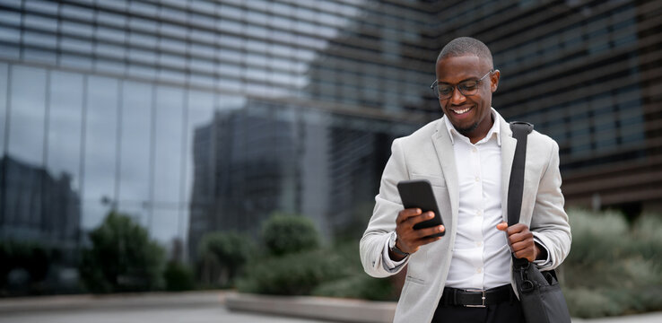Black businessman working as startup founder, using smartphone, texting, outdoor near office building. African american business man smiling, ready for next meeting, advancing. Copy space. Web banner.