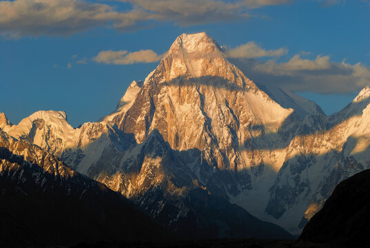 View of the snow-covered Gasherbrum IV peak illuminated by golden hour light with dramatic shadows across the rugged slopes of the Karakoram Mountain Range, Gilgit Baltistan, Pakistan.