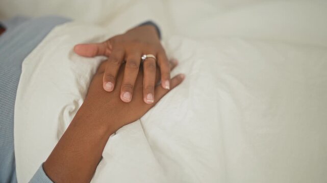 Young woman hand resting on bed with interlaced fingers and visible ring on soft white duvet in studio; calm intimacy.
