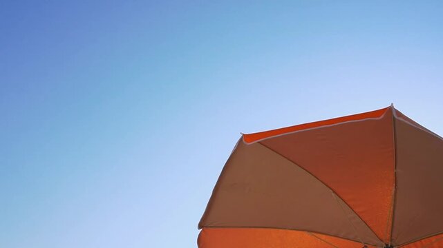 Horizontal video of a beach umbrella or sunshade view from below in the summer, seen from below, with a blue clear sky in the background, gently swaying in the wind. With copyspace to the left.