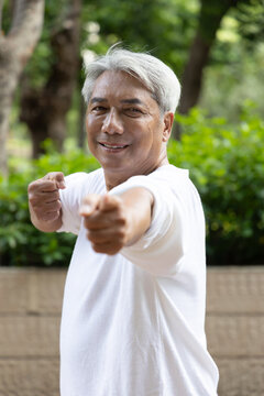 asian senior man practicing baduanjin qigong in an urban park, drawing the bow to shoot eagle pose for shoulder strength and flexibility, with relaxed smiling happy mood