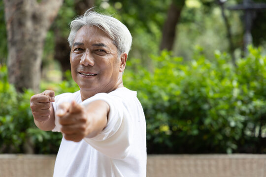 asian senior man practicing baduanjin qigong in an urban park, drawing the bow to shoot eagle pose for shoulder strength and flexibility, with relaxed smiling happy mood