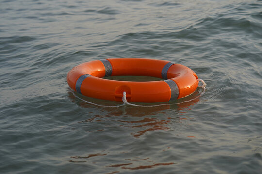 Orange life ring floats gently on calm water in ocean. Ripples surround the buoy, suggesting gentle movement. Its stripes and knot reflect safety and rescue