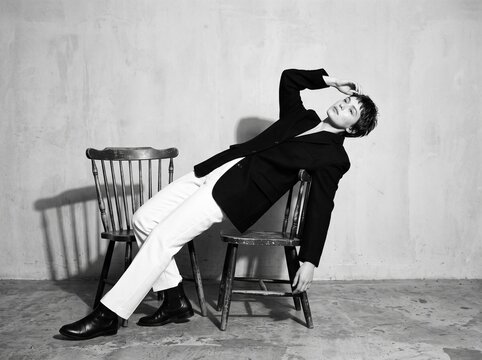 Melancholy Mood: A person draped in a black blazer and white trousers reclines on a wooden chair, with a forlorn expression and a hand touching their forehead.