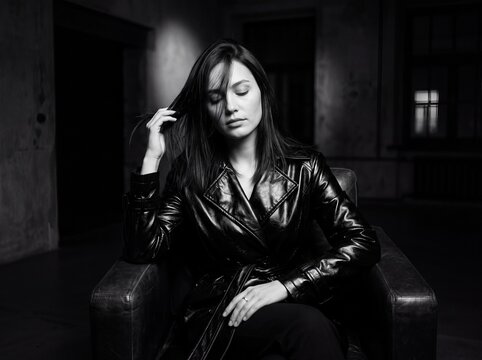 Brooding Solitude: A woman in a leather trench coat sits in a dimly lit room, her eyes closed, creating an atmosphere of introspection and melancholic beauty.