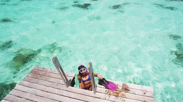 Woman after snorkel swim in paradise coral reefs ocean coming out of water removing fins to relax on overwater balcony. Raw unedited vertical video of real unretouched woman model without any makeup