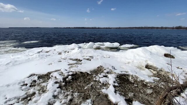 Melting ice floes and snow piled along a shoreline under a clear blue sky, bordering open dark blue river water during early spring thaw.