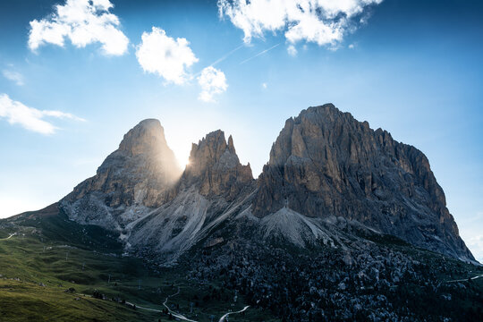 View of Sella Pass in the Dolomites with sun flare behind jagged mountain peaks and green slopes in Selva, Trentino-Alto Adige, Italy.