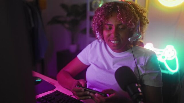 Woman with headset smiling and holding smartphone while playing and talking into a microphone at a gaming studio with neon controller sign and keyboard; joy community.
