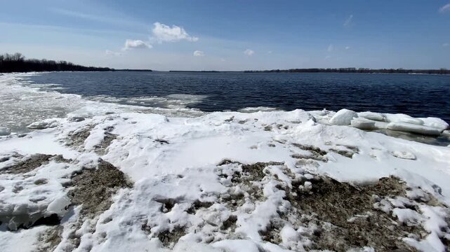 Melting ice floes and snow piled along a shoreline under a clear blue sky, bordering open dark blue river water during early spring thaw.