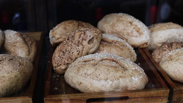 Various types of fresh, rustic, whole grain bread loaves are displayed on wooden trays inside a bakery window, showcasing traditional baked goods for sale in a warm, inviting atmosphere