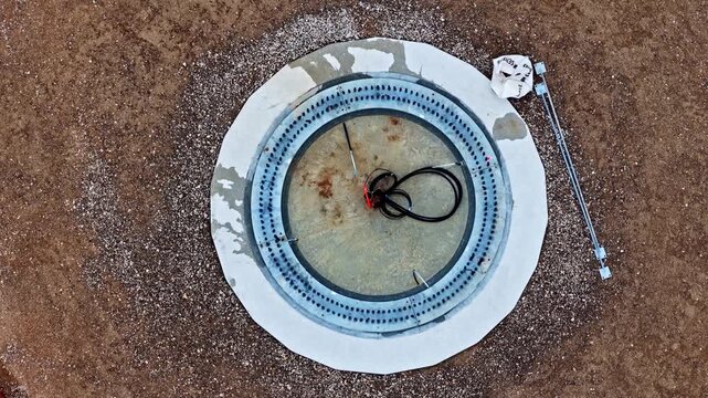 Aerial view of a circular utility hatch with exposed cables set into a textured gravel surface in Latvia.