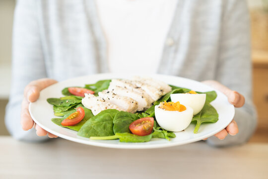 Close up of a woman holding and presenting a white plate of healthy fitness salad topped with sliced chicken breast, boiled egg and fresh tomatoes on her hand at home, weight loss meal concept.